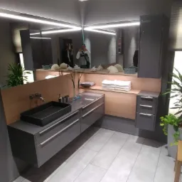 Nowoczesna łazienka w odcieniach szarości z czarną, prostokątną umywalką nablatową, podświetlanym lustrem i drewnianymi akcentami, uzupełniona o rośliny doniczkowe i drewniany stołek.