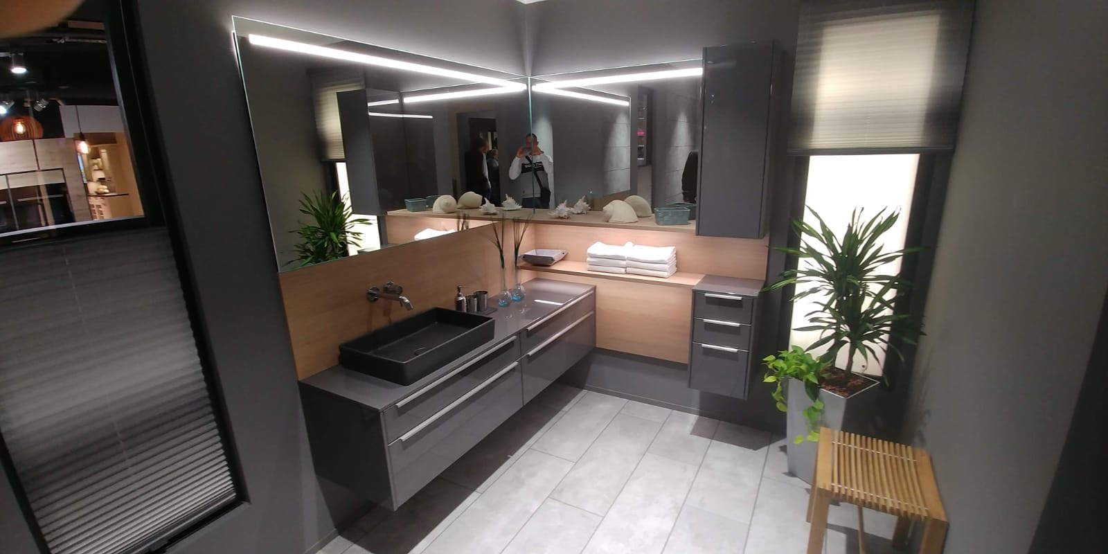 Nowoczesna łazienka w odcieniach szarości z czarną, prostokątną umywalką nablatową, podświetlanym lustrem i drewnianymi akcentami, uzupełniona o rośliny doniczkowe i drewniany stołek.