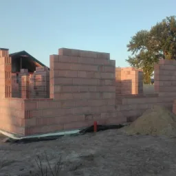 Wznoszenie ścian parterowego domu z czerwonej cegły ceramicznej, widoczne otwory okienne i drzwiowe, w tle inne budynki mieszkalne i drzewa.