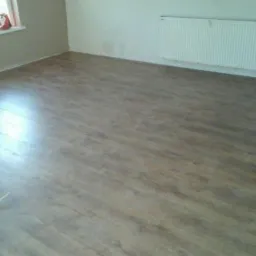 Pokój po remoncie z nową, brązową podłogą imitującą drewno, biały grzejnik na ścianie, widoczne ślady po pracach wykończeniowych.