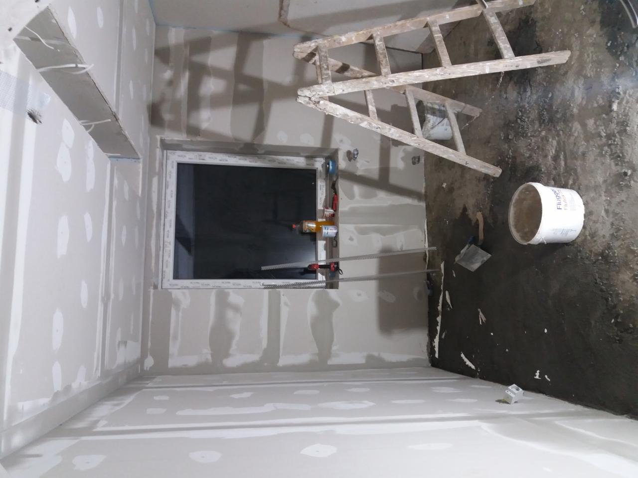 Wnętrze w Orlu w trakcie prac wykończeniowych: ściany pokryte płytami gipsowo-kartonowymi, częściowo zaszpachlowane, widoczne okno, drabina malarska i wiadro z farbą na podłodze.