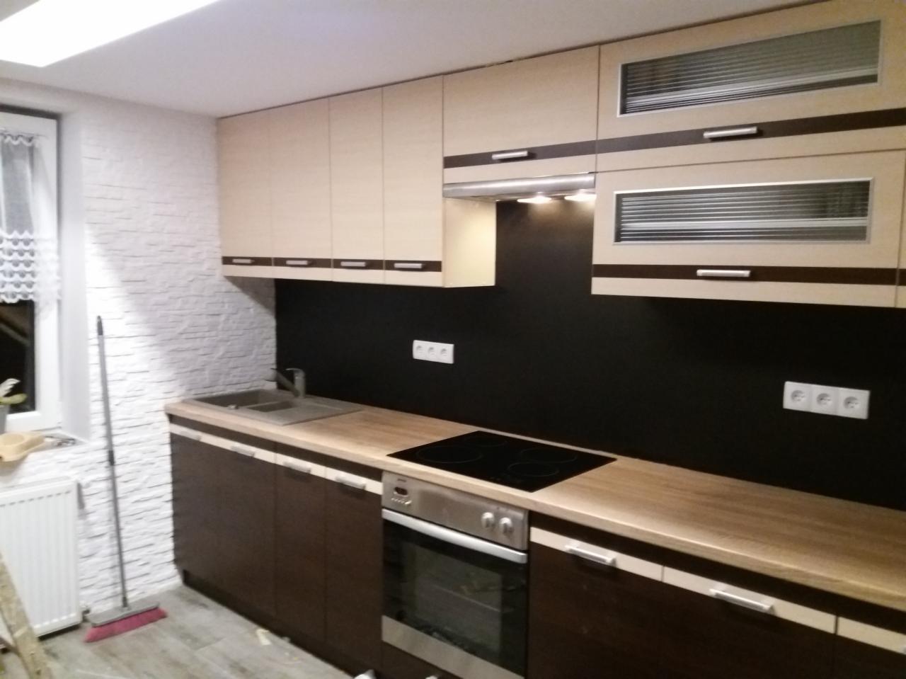 Nowoczesna kuchnia z beżowo-brązowymi szafkami, czarnym panelem nad blatem i ścianą z białej cegły dekoracyjnej. Widoczny zlew, płyta grzewcza i piekarnik. Podłoga wyłożona szarymi panelami.