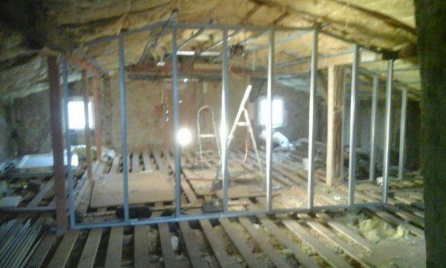 Stelaż z profili metalowych w trakcie montażu na poddaszu, widoczne drewniane deski podłogowe, izolacja na skosach dachu i drabina w tle.