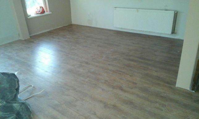 Pokój po remoncie z nową, brązową podłogą imitującą drewno, biały grzejnik na ścianie, widoczne ślady po pracach wykończeniowych.