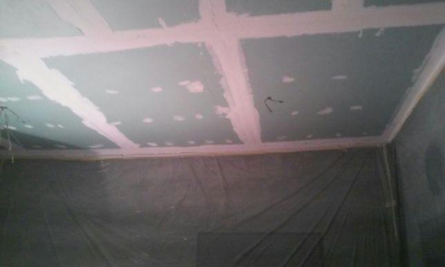 Sufit z płyt kartonowo-gipsowych w trakcie montażu, widoczne łączenia wzmocnione taśmą i pokryte szpachlą, ściany zabezpieczone folią ochronną.