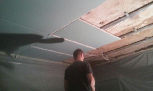 Montaż płyty karton gips na suficie, widoczne drewniane belki stropowe i częściowo przykryta folią ściana, osoba w czarnej koszulce w tle.