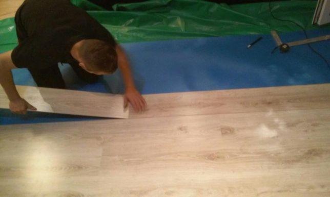 Instalacja paneli podłogowych o jasnym, drewnopodobnym wzorze; widoczne narzędzia pomiarowe i zabezpieczająca folia