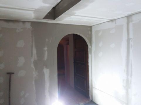 Wnętrze w trakcie szpachlowania ścian i sufitu, widoczne ślady po łatach gipsowych, łukowate przejście do innego pomieszczenia z ciemnymi drzwiami, w tle narzędzie malarskie.