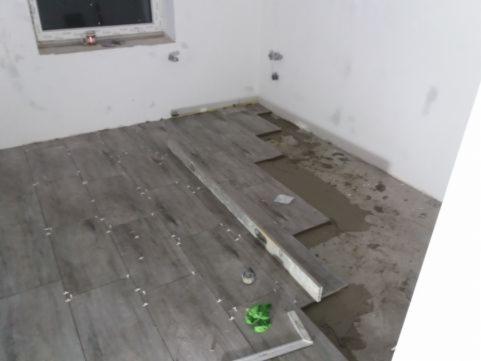 Układanie szarych płytek podłogowych w trakcie prac, widoczna poziomica i pozostałości zaprawy klejowej, zielona ścierka w rogu.