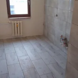 Wnętrze łazienki w trakcie remontu, z ułożonymi płytkami podłogowymi imitującymi drewno i szarymi płytkami ściennymi, widoczne wystające rury instalacji wodnej i grzejnik pod oknem.