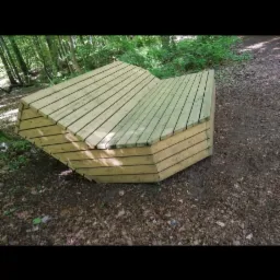 Nietypowa, wielokątna ławka z jasnego drewna, umieszczona na leśnej ścieżce pokrytej ściółką, otoczona zielenią drzew i krzewów.