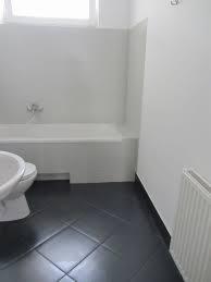 Biała łazienka z ciemną, błyszczącą podłogą z płytek ułożonych w karo. Widoczna wanna z zabudową, umywalka na postumencie i grzejnik żeberkowy. Minimalistyczny styl, brak dekoracji.