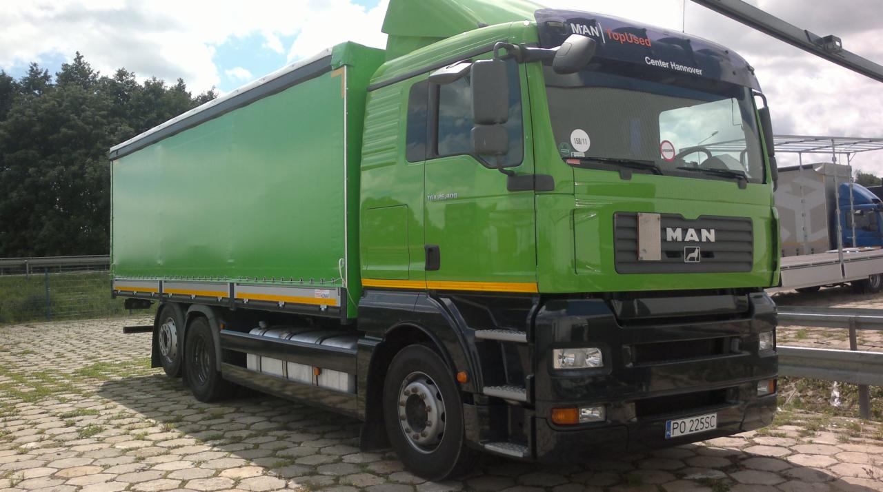 Zielona ciężarówka MAN TGS 26.400 z plandeką, zaparkowana na placu wyłożonym kostką, widoczny numer rejestracyjny i logo MAN na grillu.