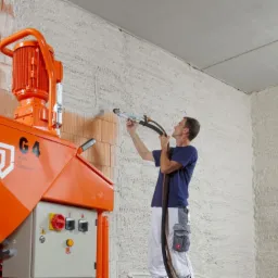 Pracownik aplikuje tynk na ścianę za pomocą pomarańczowej maszyny G4 z napisem 'MADE IN GERMANY', widoczne ślady tynku na cegłach.