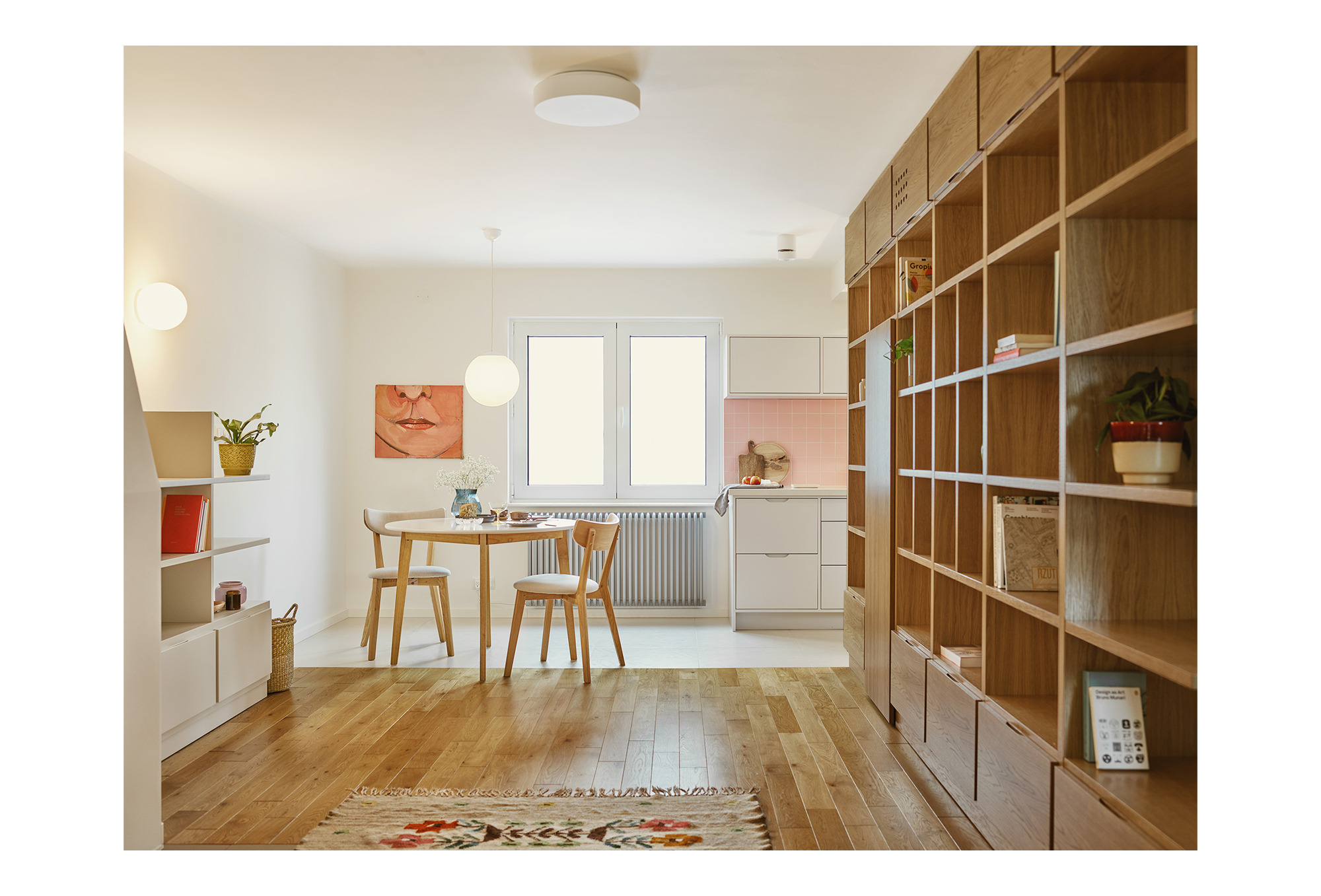 Jasne wnętrze mieszkania z drewnianą podłogą, stołem jadalnym z kwiatami, regałem z książkami i obrazem na ścianie w tle kuchni z różowymi płytkami.