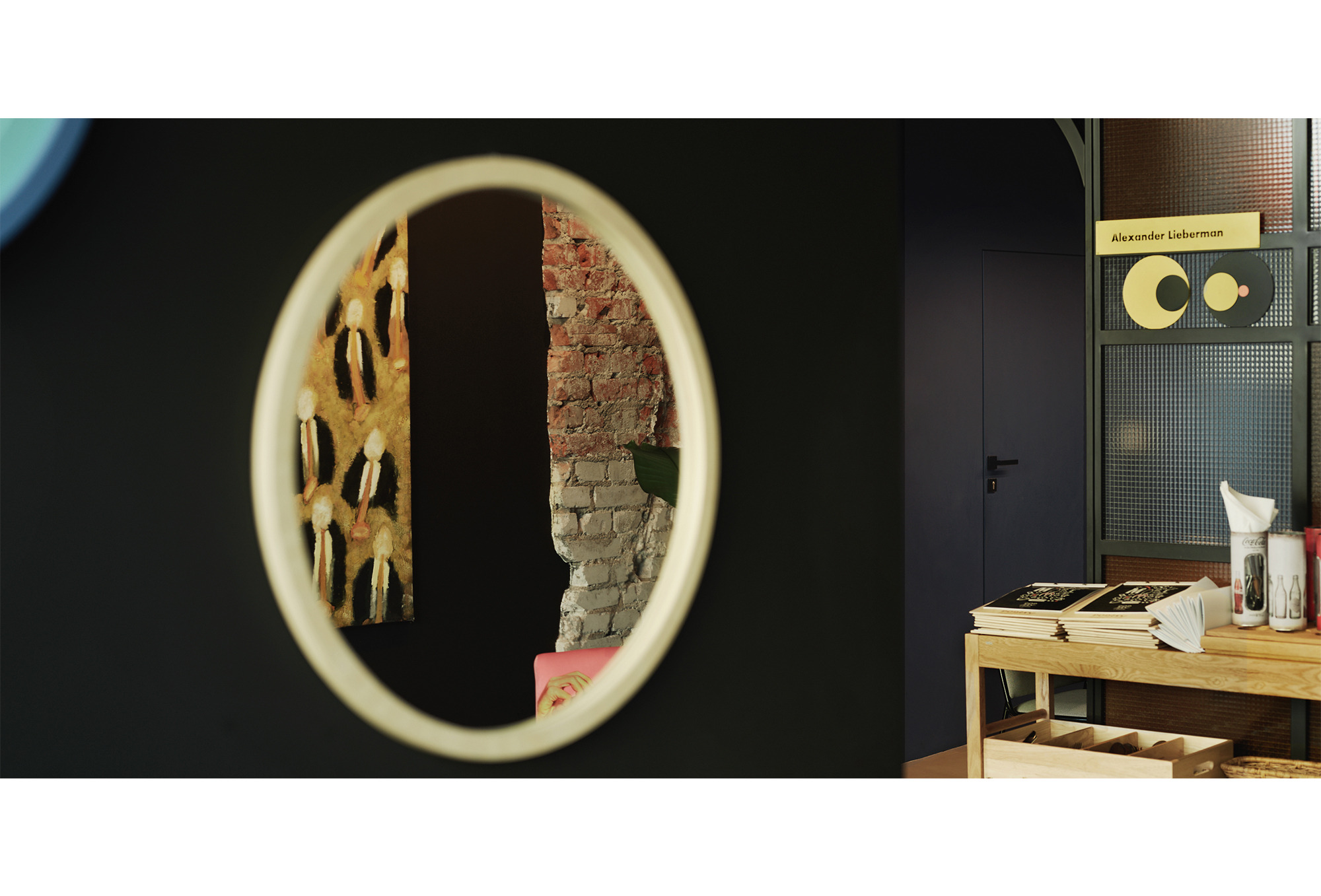 Wnętrze restauracji Pop-Inn Gdynia: okrągłe lustro odbijające ścianę z cegły i obraz z postaciami w garniturach, obok drewniany stolik z menu i napojami, na ścianie tabliczka z napisem Alexander...