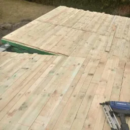 Drewniana konstrukcja dachu w trakcie budowy w Poznaniu, widoczny zszywacz pneumatyczny do mocowania desek. W tle zieleń drzew.