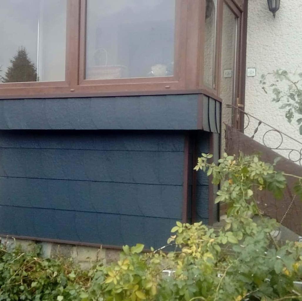 Fragment elewacji budynku z ciemnoszarą okładziną imitującą łupek, brązowe obramowania okien i narożników, widoczne schody i fragment balustrady oraz roślinność w pobliżu.