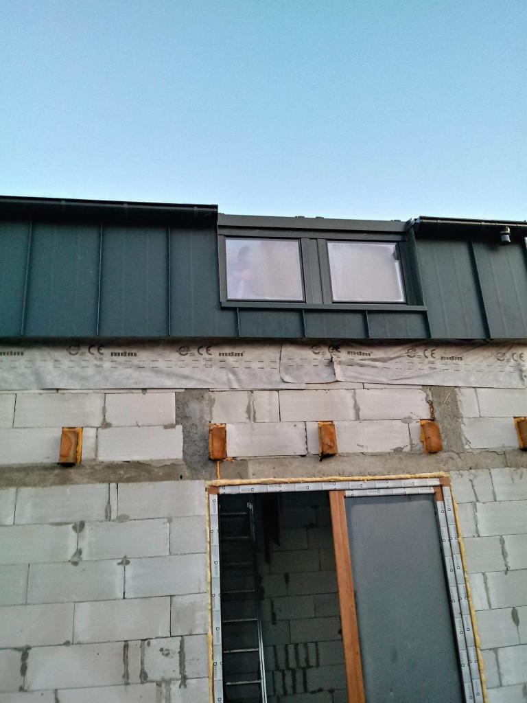 Fragment budynku w trakcie budowy: surowa ściana z betonu komórkowego, zabezpieczona folią dachową, z widocznym fragmentem dachu pokrytego blachą trapezową i oknem dachowym, na tle błękitnego nieba.