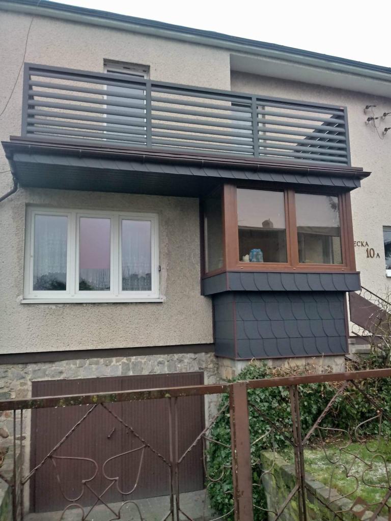 Szary budynek z balkonem z poziomymi prętami, okapem pokrytym ciemnymi płytkami imitującymi łupek i wykuszem z drewnianą ramą i podobnym pokryciem.