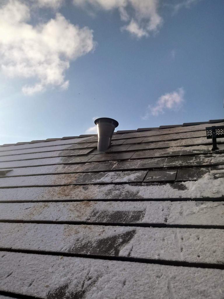 Częściowo ośnieżony dach z szarej dachówki z kominem wentylacyjnym na tle błękitnego nieba z chmurami.