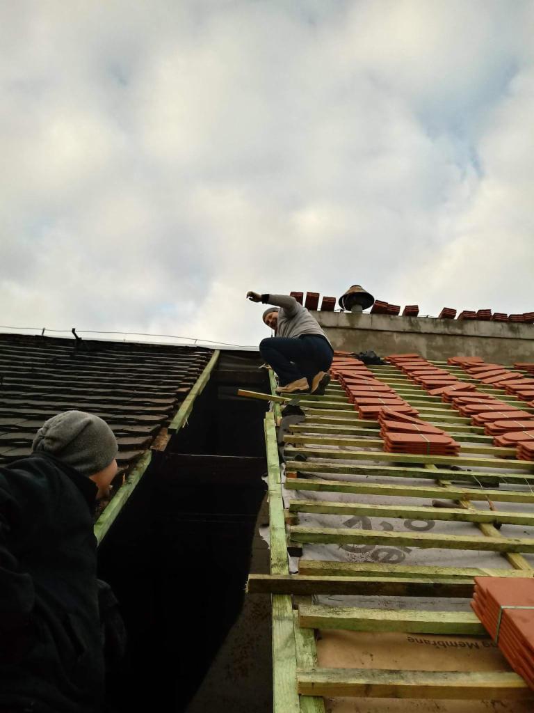 Dekarze pracują nad wymianą dachu: częściowo zdjęte stare dachówki, widoczna membrana, nowe dachówki ułożone w rzędach, pochmurne niebo w tle.