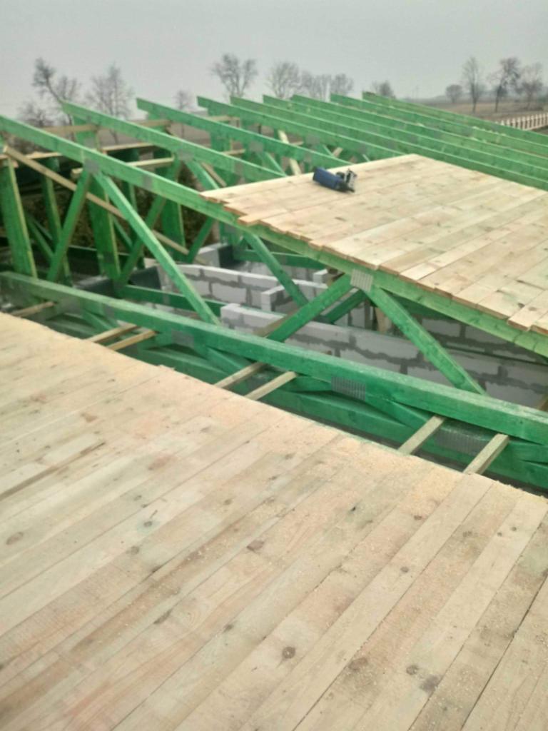 Konstrukcja drewniana dachu z zielonymi belkami, częściowo pokryta deskami, widoczne bloczki betonowe i narzędzie na dachu, w tle drzewa.