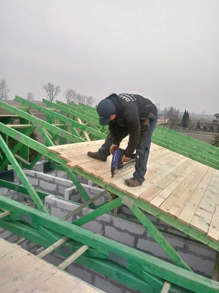 Pracownik w trakcie montażu desek na zielonej konstrukcji więźby dachowej za pomocą gwoździarki pneumatycznej, widoczne bloczki betonowe i ponura pogoda w tle.