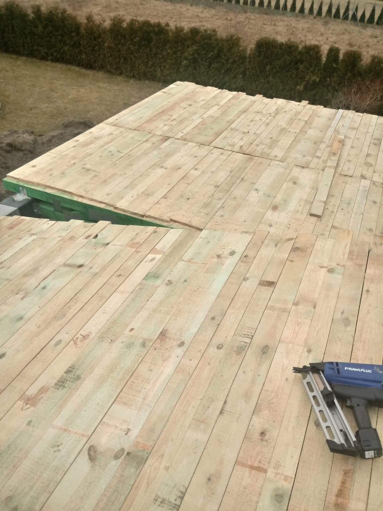 Drewniana konstrukcja dachu w trakcie budowy w Poznaniu, widoczny zszywacz pneumatyczny do mocowania desek. W tle zieleń drzew.