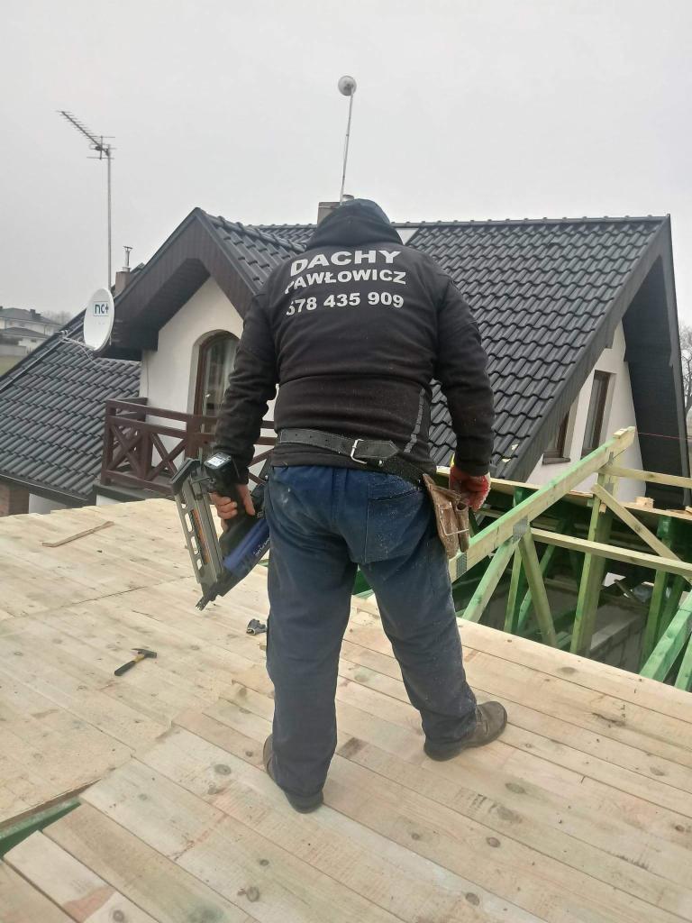 Dekarz w czarnej bluzie z napisem 'DACHY PAWŁOWICZ 578 435 909' stojący na drewnianej konstrukcji dachu z narzędziem do wbijania gwoździ w ręku, w tle budynek z ciemnym dachem i antenami.