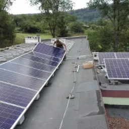 Instalator umieszcza panel słoneczny na metalowej konstrukcji dachu budynku, w tle widoczne wzgórza i drzewa.