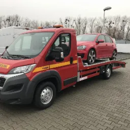 Czerwona laweta z napisem 'Pomoc Drogowa' przewożąca czerwony samoch&oacute;d osobowy na tle szarej kostki brukowej i drzew bez liści.