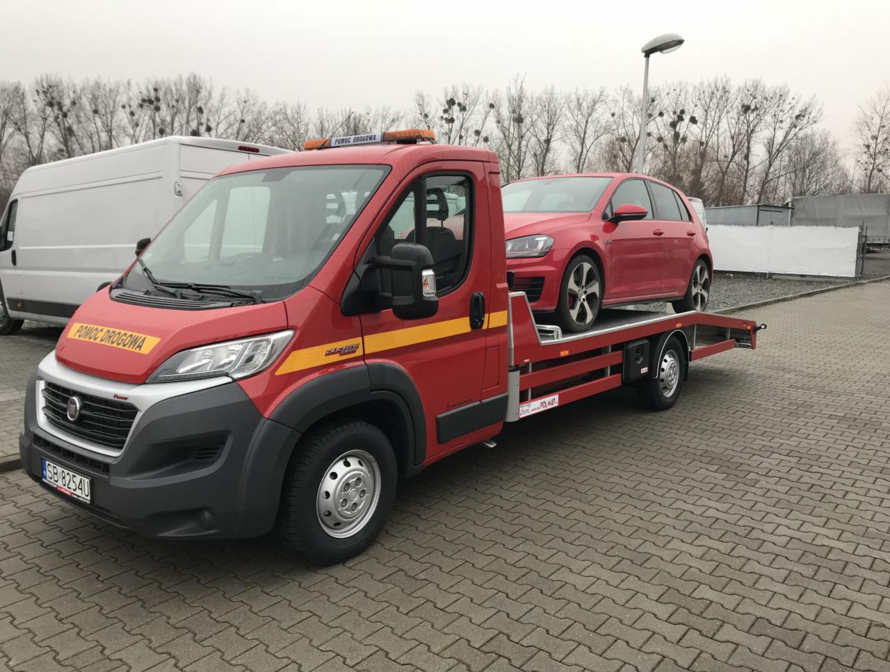 Czerwona laweta z napisem 'Pomoc Drogowa' przewożąca czerwony samochód osobowy na tle szarej kostki brukowej i drzew bez liści.