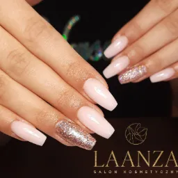 Dłonie z jasnoróżowym, kwadratowym manicure, jeden paznokieć każdej dłoni pokryty brokatem, widoczne logo salonu kosmetycznego w tle.