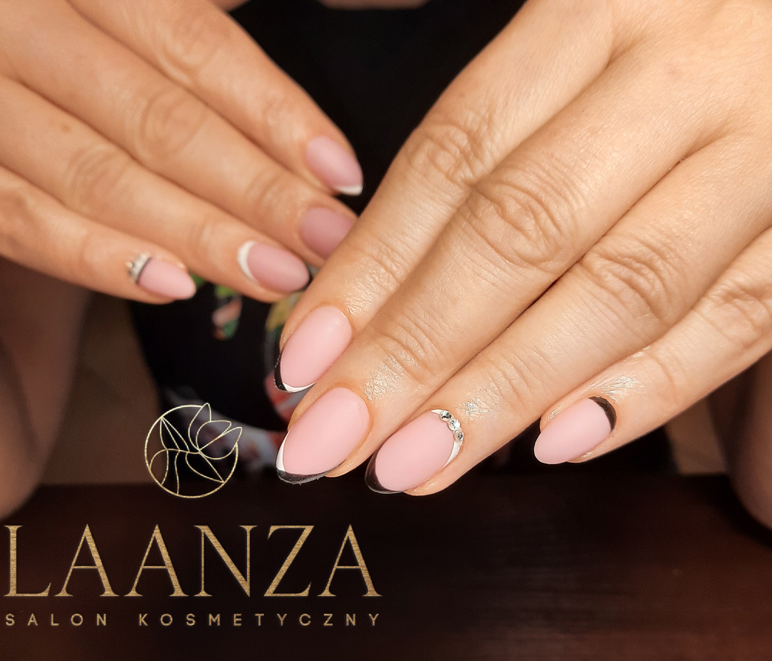 Dłonie z manicure hybrydowym w odcieniach różu i czerni, z delikatnymi zdobieniami w postaci białych pasków i cyrkonii, na tle wizytówki salonu kosmetycznego Laanza.