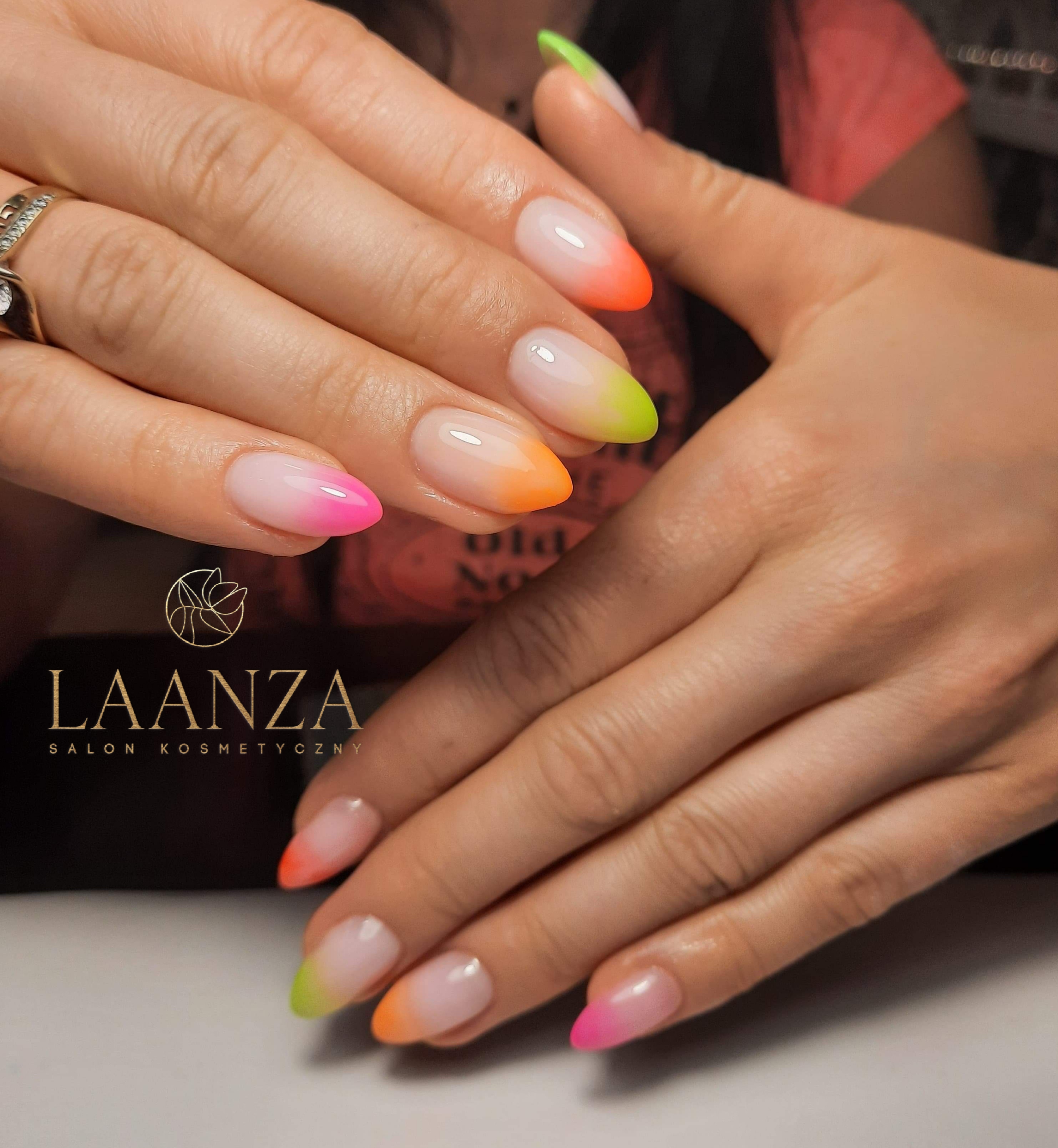 Dłonie z owalnymi paznokciami pomalowanymi w stylu ombre, przechodzącymi od jasnego różu, pomarańczu i limonki na końcach, na tle logo salonu kosmetycznego Laanza.