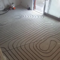Wykonywanie instalacji podłogowej z rur grzewczych w surowym pomieszczeniu, widoczny rozkład pętli i szlifierka do betonu w tle.