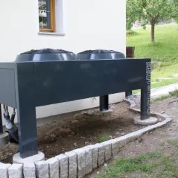 Szara pompa ciepła z dwoma wentylatorami, zamontowana na betonowych fundamentach otoczonych kamiennym obrzeżem, widoczna instalacja rurowa, trawnik w tle.