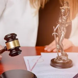 Kobieta w białym żakiecie trzyma młotek sędziowski nad dokumentami, obok stoi statuetka Temidy na drewnianym biurku.