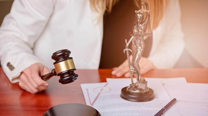 Kobieta w białym żakiecie trzyma młotek sędziowski nad dokumentami, obok stoi statuetka Temidy na drewnianym biurku.