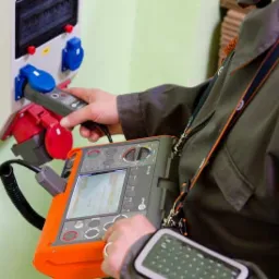 Osoba w kombinezonie roboczym wykonuje pomiary gniazd przemysłowych za pomocą miernika elektrycznego z pomarańczową obudową i wyświetlaczem LCD.