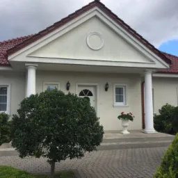 Jasna elewacja domu jednorodzinnego z kolumnami, czerwoną dachówką i zadbanym ogrodem z kostki brukowej.