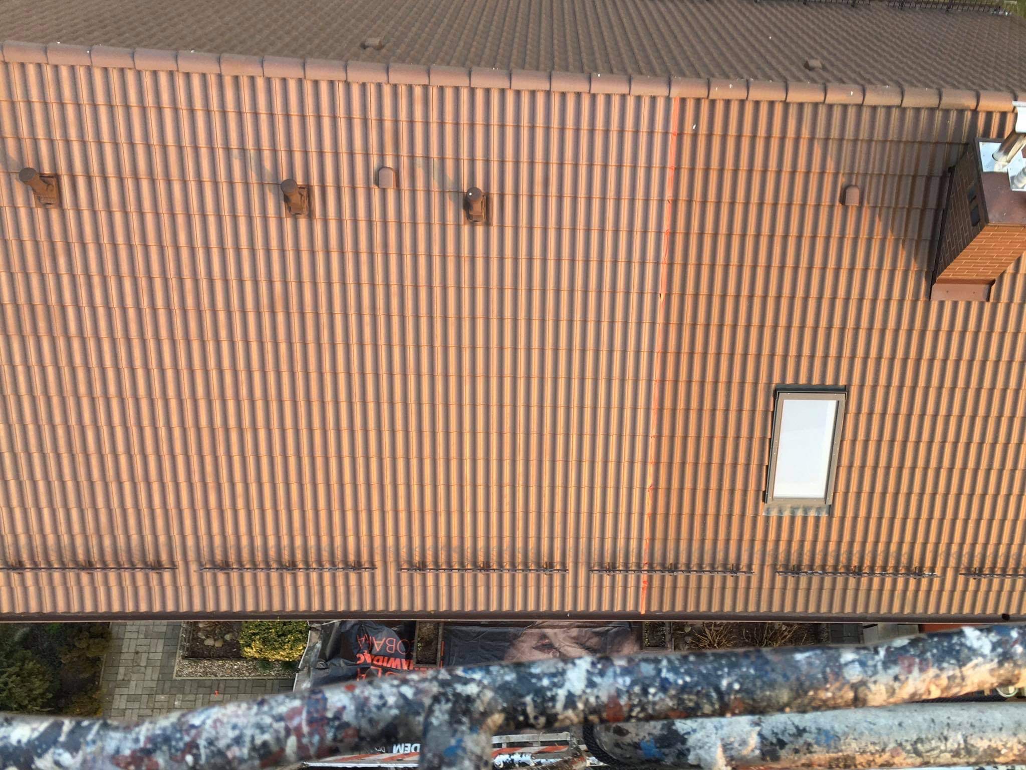 Widok z góry na brązowy dach pokryty dachówką ceramiczną, z widocznym oknem dachowym i elementami wentylacji, sfotografowany z rusztowania z widocznym fragmentem ogrodu poniżej.
