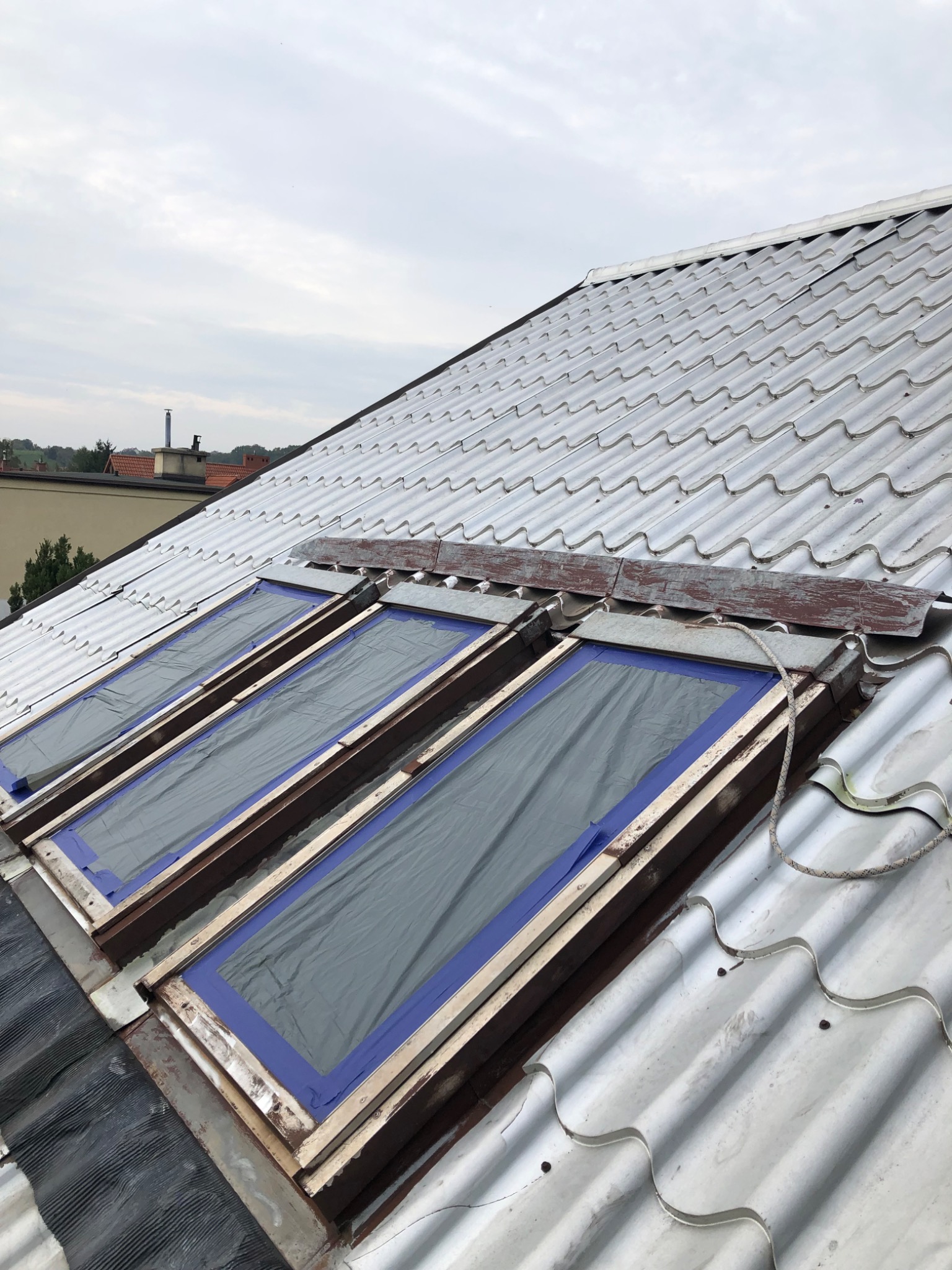 Biała dachówka falista z trzema oknami dachowymi zabezpieczonymi folią i taśmą malarską, widoczne ślady korozji na metalowych elementach.