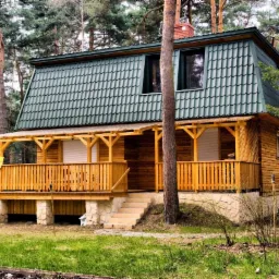 Drewniany dom z zielonym dachem dwuspadowym, widoczny z bliskiej perspektywy, otoczony wysokimi drzewami iglastymi, z werandą i kamienną podmurówką.