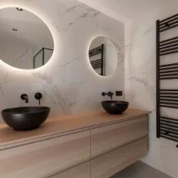 Nowoczesna łazienka z dwoma czarnymi umywalkami nablatowymi, podświetlanymi okrągłymi lustrami, drewnianą szafką z szufladami i czarnym grzejnikiem drabinkowym na tle marmurowych płytek.