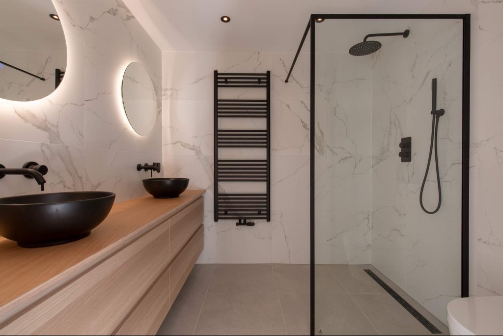 Nowoczesna łazienka z dwoma czarnymi umywalkami nablatowymi, drewnianą szafką pod umywalkami, podświetlanymi okrągłymi lustrami, czarnym grzejnikiem drabinkowym i kabiną prysznicową z czarnymi...