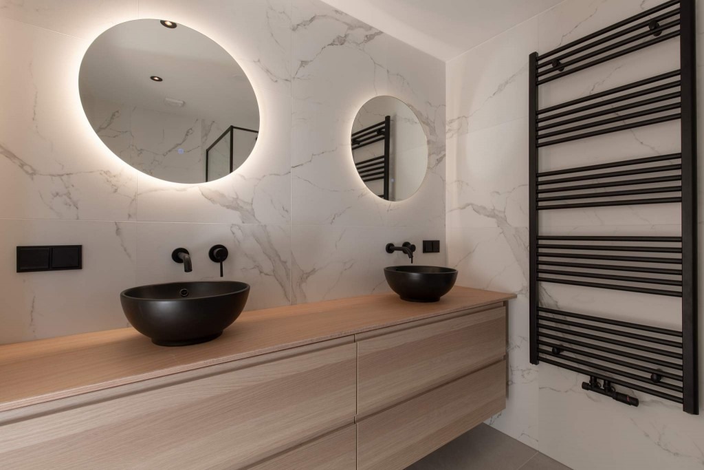 Nowoczesna łazienka z dwoma czarnymi umywalkami nablatowymi, podświetlanymi okrągłymi lustrami i grzejnikiem drabinkowym w kolorze czarnym na tle białych płytek imitujących marmur.