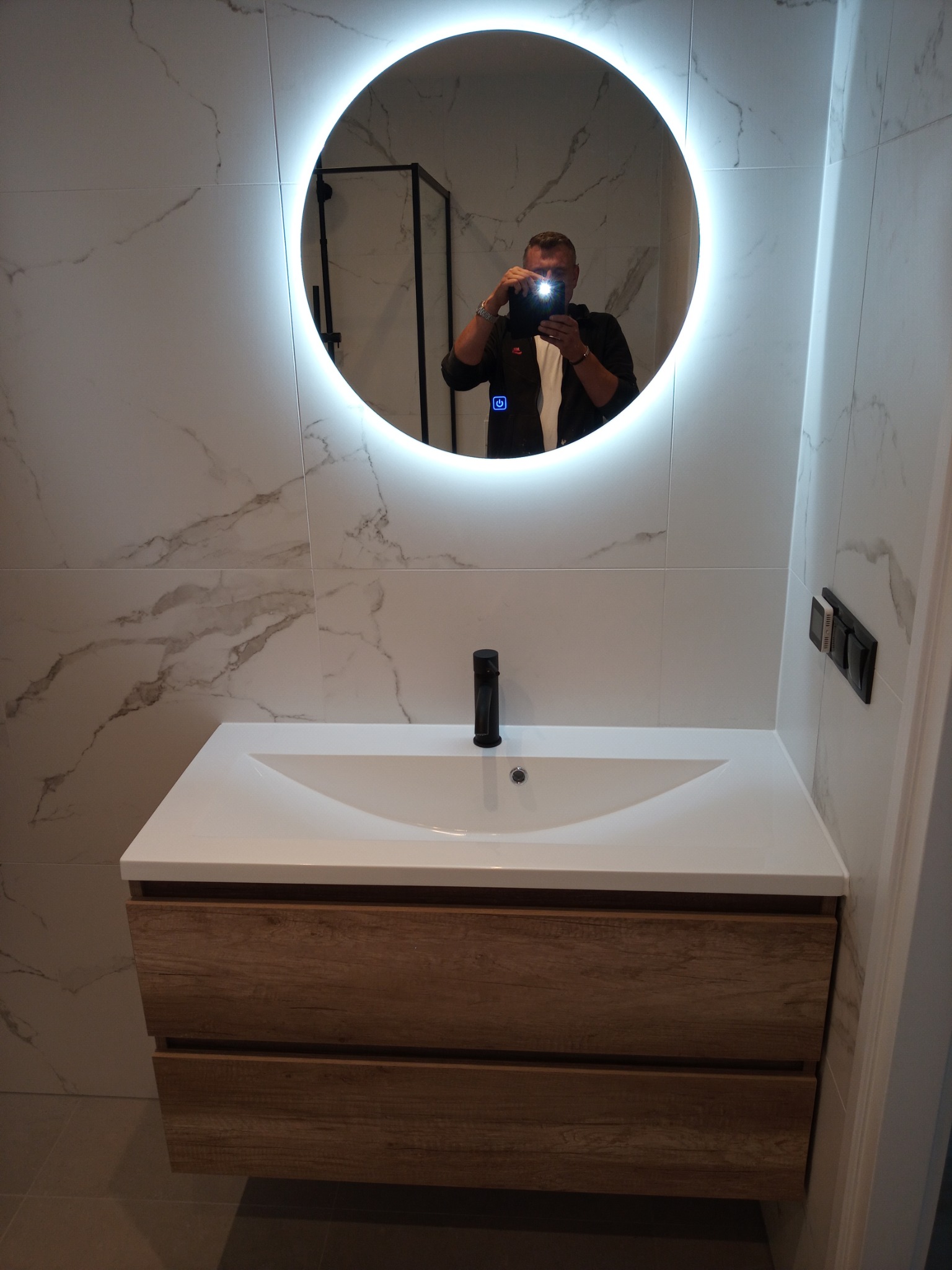 Nowoczesna łazienka z umywalką nablatową na szafce z drewnopodobnym frontem, okrągłe lustro z podświetleniem LED, marmurowe płytki na ścianach, widoczne odbicie osoby robiącej zdjęcie.