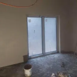 Świeżo otynkowane wnętrze z oknem zabezpieczonym folią, widoczne narzędzia i wiadro po tynku, niedokończone prace wykończeniowe.